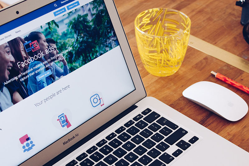 Laptop open on a table with a page about Facebook ads pulled up in a browser; next to the computer is a glass, a mouse, and a pen