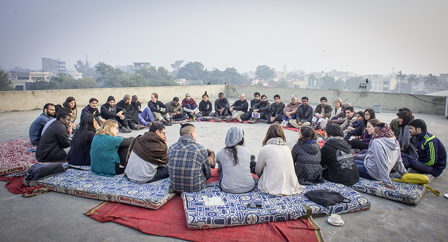 a group of people sitting in a circle on cushions outside discussing something