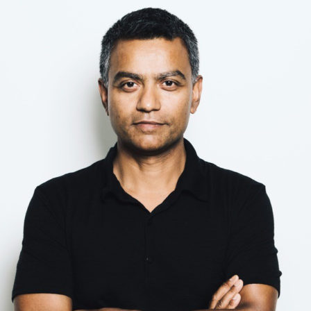 Deb Roy - a medium brown skinned man with short black hair grey at the temples wearing a black polo Tshirt, his arms crossed, looking at the camera with a subtle smile