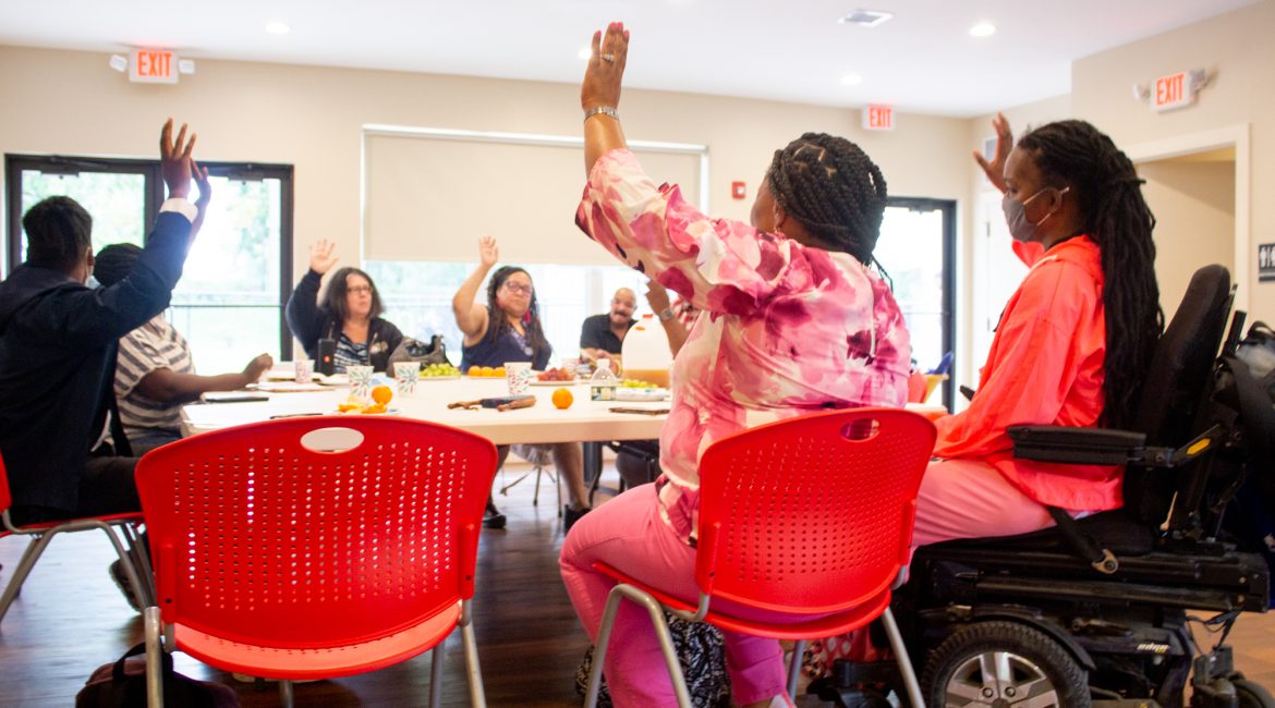 A group of people - most of whom are Black or African American - sitting around a table in red chairs with their hands raised.