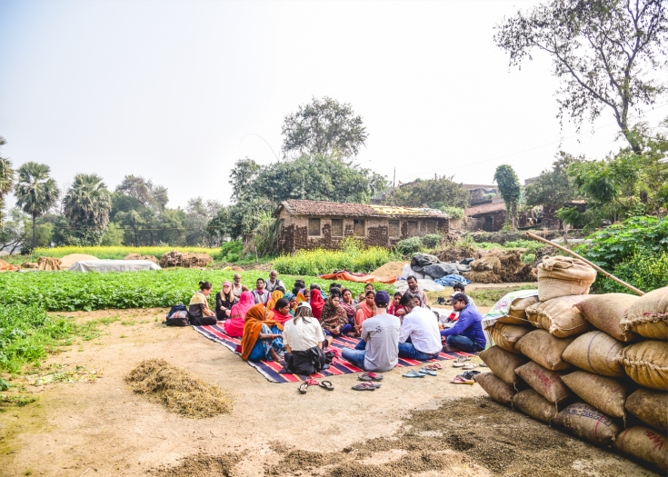 A group of people sitting outside in a circle on brightly colored mats in a village. There is a garden and small house behind them and tall tropical trees in the background. To the right is a stack of many bags of harvested items.